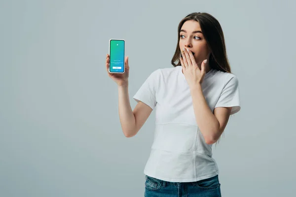 KYIV, UCRANIA - 6 de junio de 2019: impactada hermosa chica en camiseta blanca que muestra el teléfono inteligente con la aplicación de twitter aislada en gris - foto de stock