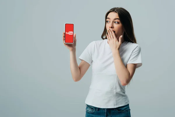 КИЕВ, Украина - 6 июня 2019 года: шокированная красивая девушка в белой футболке, показывающая смартфон с приложением youtube, изолированным на сером — стоковое фото