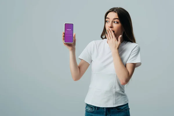 KYIV, UCRANIA - 6 de junio de 2019: impactada hermosa chica en camiseta blanca que muestra el teléfono inteligente con aplicación de Instagram aislado en gris - foto de stock