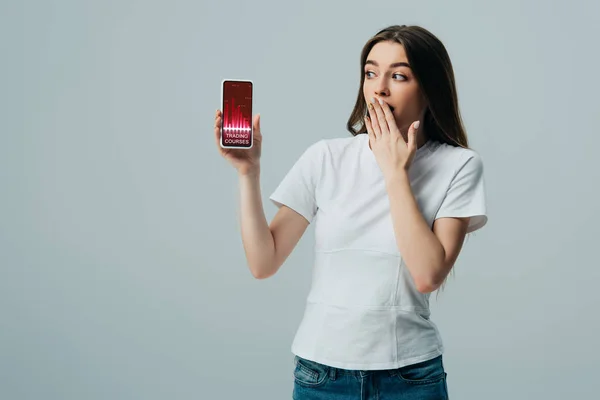 Impactado hermosa chica en camiseta blanca que muestra el teléfono inteligente con la aplicación de cursos de comercio aislado en gris - foto de stock