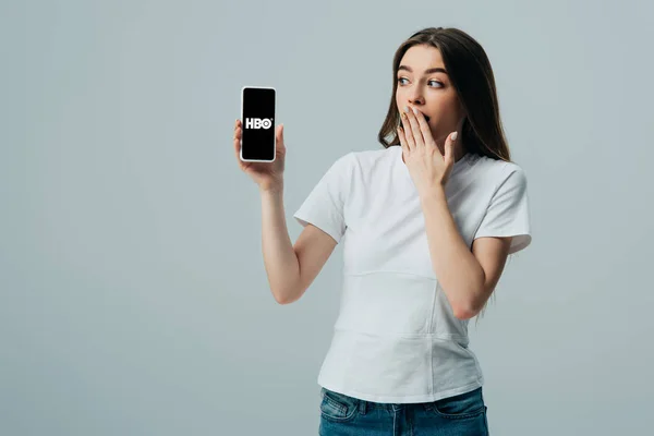 KYIV, UCRANIA - 6 de junio de 2019: impactada hermosa chica en camiseta blanca que muestra el teléfono inteligente con la aplicación HBO aislada en gris - foto de stock