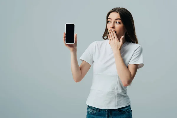Шокована красива дівчина в білій футболці, що показує смартфон з порожнім екраном ізольовано на сірому — стокове фото