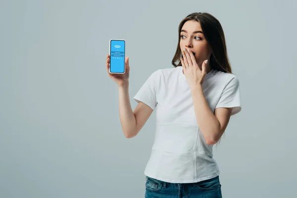 KYIV, UKRAINE - JUNHO 6, 2019: Menina bonita chocada em camiseta branca mostrando smartphone com aplicativo Skype isolado em cinza — Fotografia de Stock