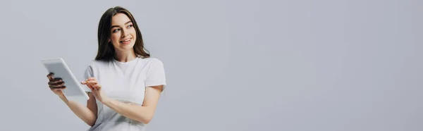 Щаслива дівчина в білій футболці тримає цифровий планшет і дивиться ізольовано на сірий, панорамний постріл — стокове фото