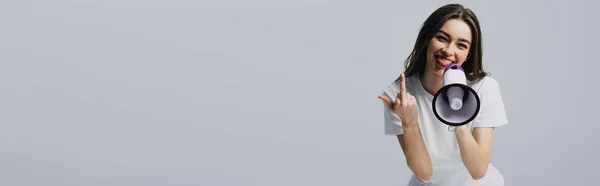 Счастливая молодая симпатичная женщина с громкоговорителем, показывающим средний палец на сером, панорамном снимке — стоковое фото