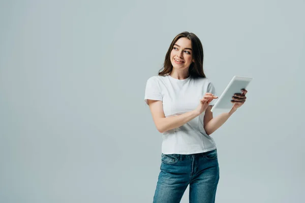 Glückliches Mädchen im weißen T-Shirt, das ein digitales Tablet in der Hand hält und isoliert auf grau blickt — Stockfoto