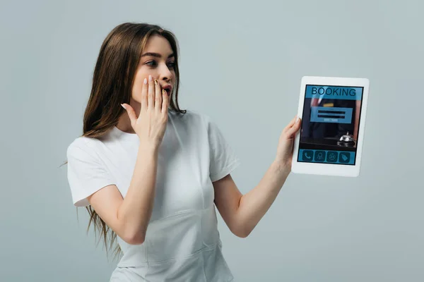 Shocked beautiful girl in white t-shirt showing digital tablet with booking app isolated on grey — Stock Photo