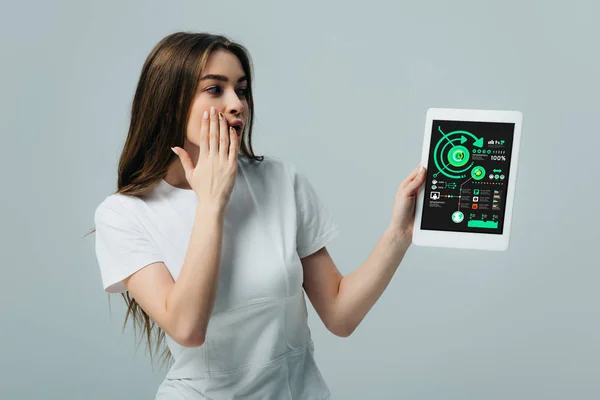 Impactado hermosa chica en camiseta blanca que muestra tableta digital con aplicación de infografías aisladas en gris - foto de stock