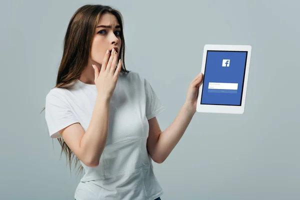 KYIV, UKRAINE - 6 JUIN 2019 : belle fille choquée en t-shirt blanc montrant tablette numérique avec application facebook isolée sur gris — Photo de stock