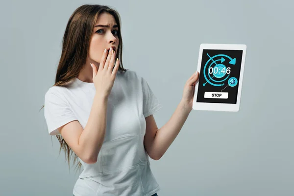 Impactado hermosa chica en camiseta blanca que muestra la tableta digital con la aplicación de cronómetro aislado en gris - foto de stock