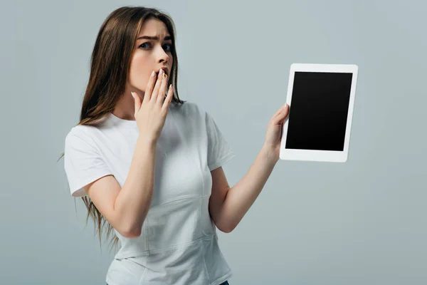 Impactado hermosa chica en camiseta blanca que muestra tableta digital con pantalla en blanco aislado en gris - foto de stock