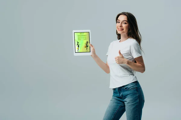 Lächelndes schönes Mädchen in weißem T-Shirt zeigt digitales Tablet mit Online-Shopping-App und zeigt Daumen hoch vereinzelt auf grau — Stockfoto