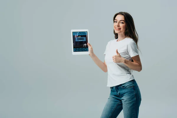 Sonriente hermosa chica en camiseta blanca mostrando tableta digital con aplicación de reserva en línea y mostrando el pulgar hacia arriba aislado en gris - foto de stock