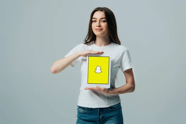 KYIV, UKRAINE - 6 JUIN 2019 : belle fille souriante en t-shirt blanc montrant tablette numérique avec application Snapchat isolée sur gris — Photo de stock