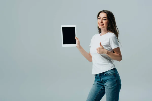 Lächelndes schönes Mädchen in weißem T-Shirt zeigt digitales Tablet mit leerem Bildschirm und Daumen nach oben isoliert auf grau — Stockfoto