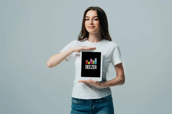 КИЕВ, УКРАИНА - 6 ИЮНЯ 2019 года: улыбающаяся красивая девушка в белой футболке с цифровым планшетом с приложением Deezer, изолированным на сером — стоковое фото