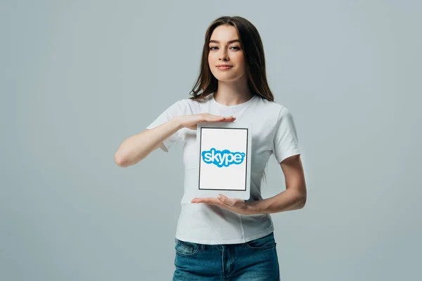 KYIV, UCRANIA - 6 de junio de 2019: hermosa niña sonriente en camiseta blanca que muestra la tableta digital con la aplicación de Skype aislada en gris — Stock Photo
