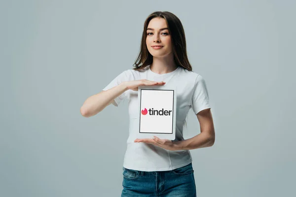 KYIV, UKRAINE - JUNE 6, 2019: smiling beautiful girl in white t-shirt showing digital tablet with Tinder app isolated on grey — Stock Photo