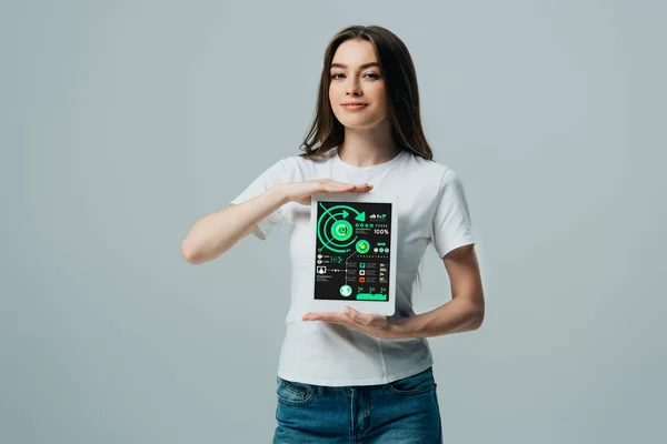 Улыбающаяся красивая девушка в белой футболке, показывающая цифровой планшет с инфографическим приложением, изолированным на сером — стоковое фото