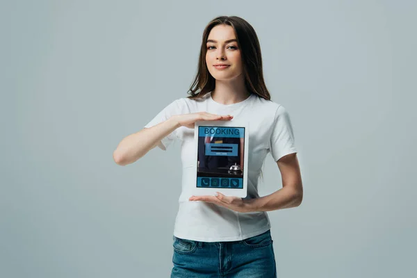 Sonrisa hermosa chica en camiseta blanca mostrando tableta digital con aplicación de reserva aislado en gris - foto de stock