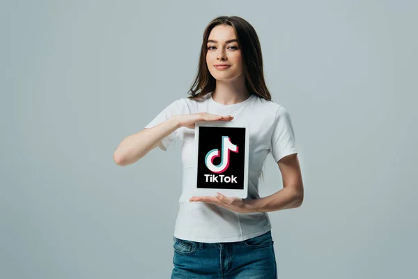 KYIV, UCRANIA - 6 de junio de 2019: hermosa niña sonriente en camiseta blanca que muestra la tableta digital con la aplicación Tik Tok aislada en gris — Stock Photo