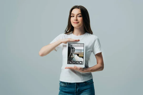 Улыбающаяся красивая девушка в белой футболке показывает цифровой планшет с билетами онлайн приложение изолированы на серый — стоковое фото