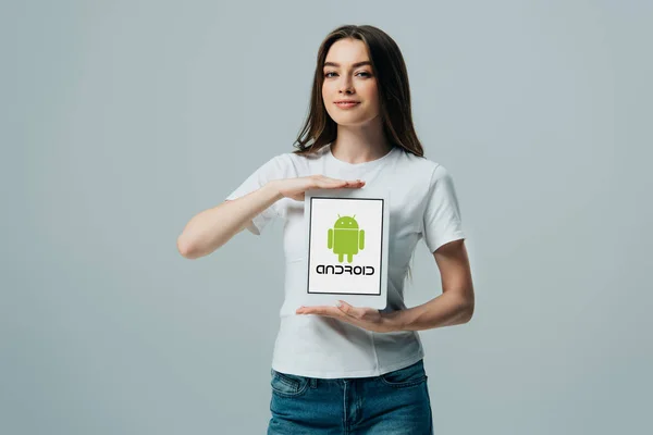КИЕВ, Украина - 6 июня 2019 года: улыбающаяся красивая девушка в белой футболке с цифровым планшетом с иконкой Android, изолированной на сером — стоковое фото