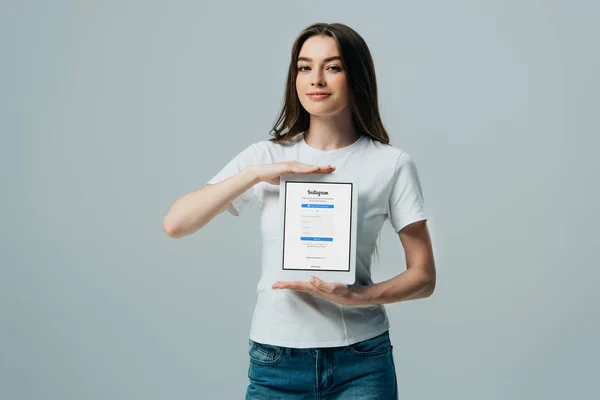 КИЕВ, УКРАИНА - 6 июня 2019 года: улыбающаяся красивая девушка в белой футболке с цифровым планшетом с приложением Instagram, изолированным на сером — стоковое фото
