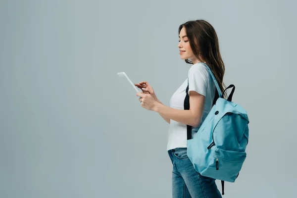 Side view of pretty girl with blue backpack using digital tablet isolated on grey — Stock Photo