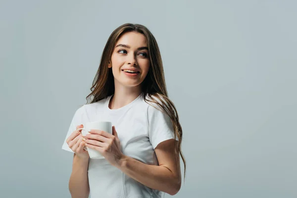 Chica feliz en camiseta blanca sosteniendo taza blanca y mirando hacia otro lado aislado en gris - foto de stock