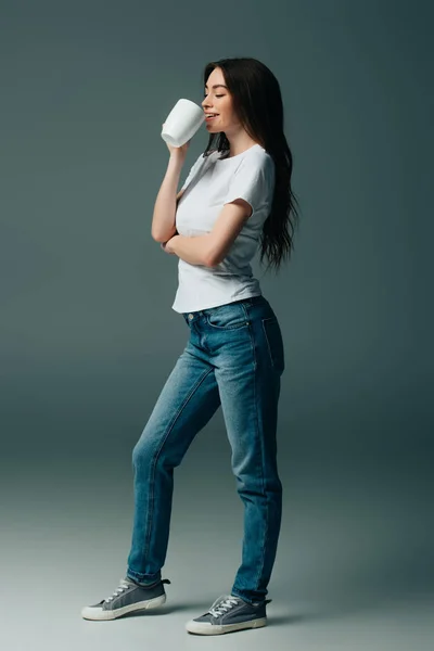 Вид в полный рост улыбающейся девушки в белой футболке и джинсах, держащей белую кружку — стоковое фото