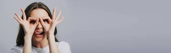 Panoramic shot of beautiful happy girl showing tongue and ok signs isolated on grey — Stock Photo