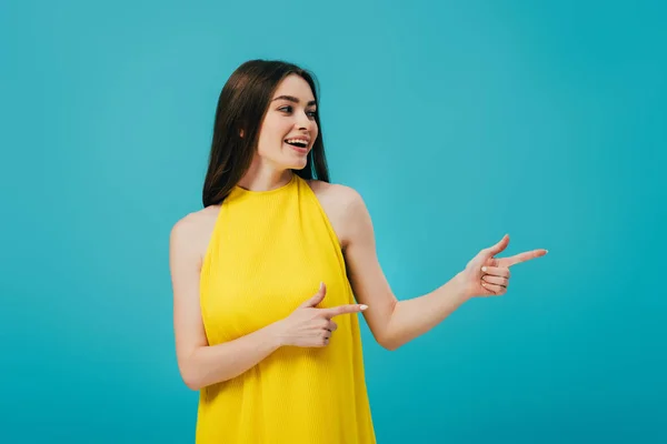 Feliz hermosa chica en vestido amarillo señalando con los dedos y mirando hacia otro lado aislado en turquesa - foto de stock