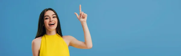 Feliz hermosa chica en vestido amarillo apuntando con el dedo hacia arriba aislado en azul, tiro panorámico - foto de stock