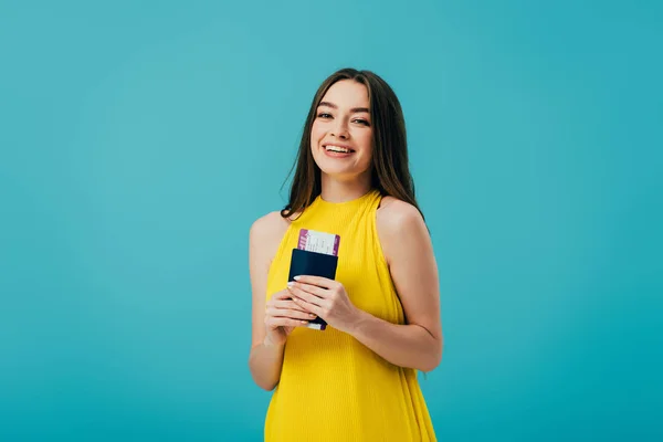 Happy brunette girl in yellow dress holding passport with air ticket isolated on turquoise — Stock Photo