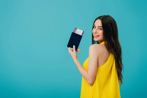 Back view of happy brunette woman in yellow dress holding passport with air ticket isolated on turquoise — Stock Photo