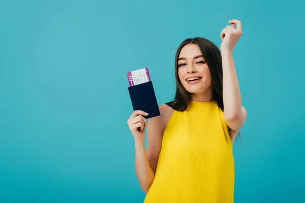 Весела брюнетка в жовтій сукні танцює з паспортом з авіаквитком ізольована на бірюзовому — стокове фото