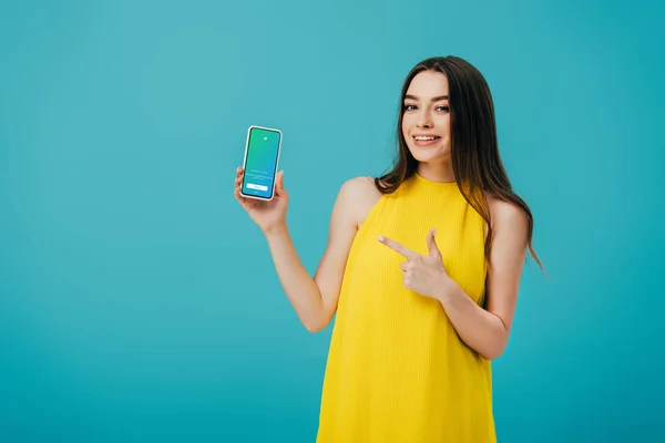 KYIV, UCRANIA - 6 de junio de 2019: hermosa chica feliz en vestido amarillo apuntando con el dedo al teléfono inteligente con la aplicación de twitter aislada en turquesa - foto de stock