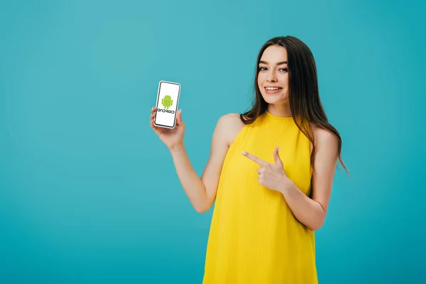 KYIV, UCRANIA - 6 de junio de 2019: hermosa niña feliz en vestido amarillo que apunta con el dedo al teléfono inteligente con el logotipo de Android aislado en turquesa - foto de stock