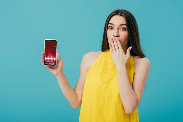 Schockiertes schönes Mädchen in gelbem Kleid zeigt Smartphone mit Trading-App isoliert auf Türkis — Stockfoto