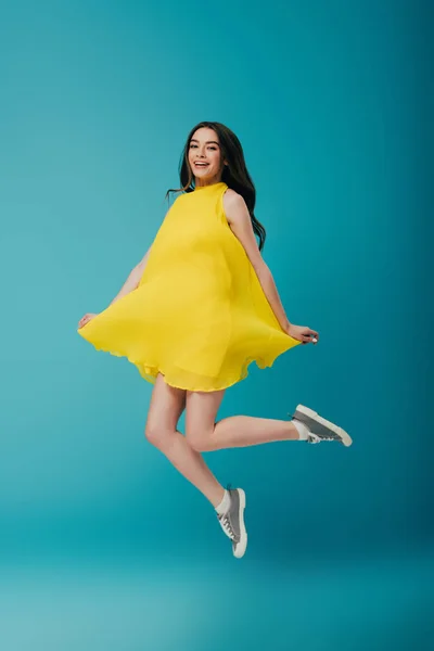 Full length view of happy girl in yellow dress jumping on turquoise background — Stock Photo