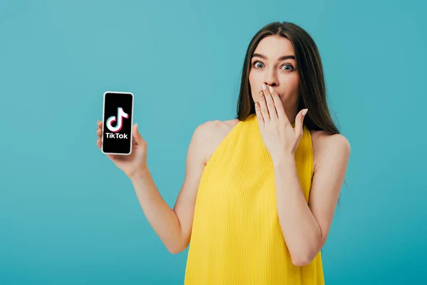 KYIV, UCRANIA - 6 de junio de 2019: impactada hermosa chica en vestido amarillo que muestra el teléfono inteligente con la aplicación Tik Tok aislada en turquesa - foto de stock