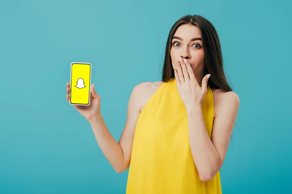 KYIV, UCRANIA - 6 de junio de 2019: impactada hermosa chica en vestido amarillo que muestra el teléfono inteligente con la aplicación Snapchat aislado en turquesa - foto de stock