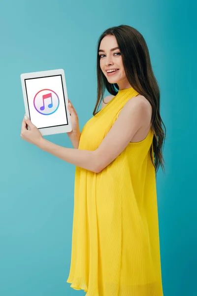 KYIV, UCRAINA - 6 GIUGNO 2019: bella ragazza sorridente in abito giallo che mostra tablet digitale con app iTunes isolata su turchese — Foto stock