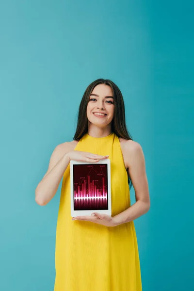Linda menina morena em vestido amarelo mostrando tablet digital com aplicativo de negociação isolado em turquesa — Fotografia de Stock