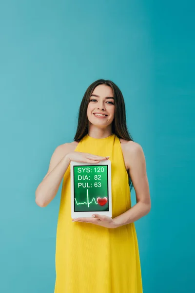 Belle fille brune en robe jaune montrant tablette numérique avec application cardiologique isolée sur turquoise — Photo de stock