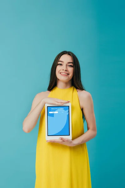 KYIV, UCRANIA - 6 de junio de 2019: hermosa niña sonriente en vestido amarillo mostrando tableta digital con aplicación de twitter aislada en turquesa - foto de stock