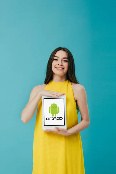 КИЕВ, УКРАИНА - 6 ИЮНЯ 2019 года: улыбающаяся красивая девушка в желтом платье с цифровым планшетом с логотипом андроида, изолированным на бирюзовом — стоковое фото