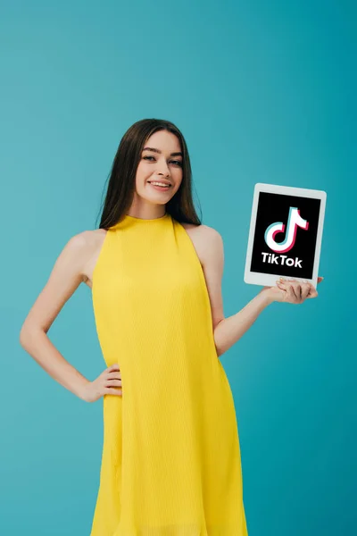 KYIV, UKRAINE - JUNE 6, 2019: smiling girl in yellow dress with hand on hip showing digital tablet with app isolated on turquoise — Stock Photo