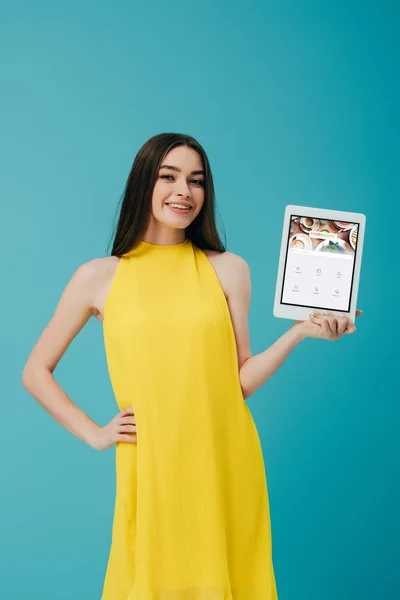 KYIV, UCRAINA - 6 GIUGNO 2019: ragazza sorridente in abito giallo con mano sull'anca che mostra tablet digitale con app quadrata isolata su turchese — Foto stock